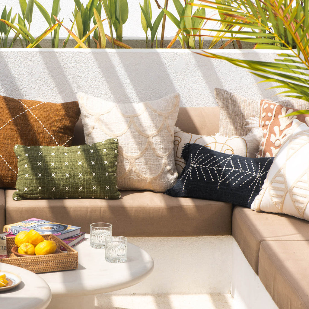 Outdoor seating area with a beige sectional sofa adorned with various throw pillows, a small table with fruit and glasses, and greenery in the background.