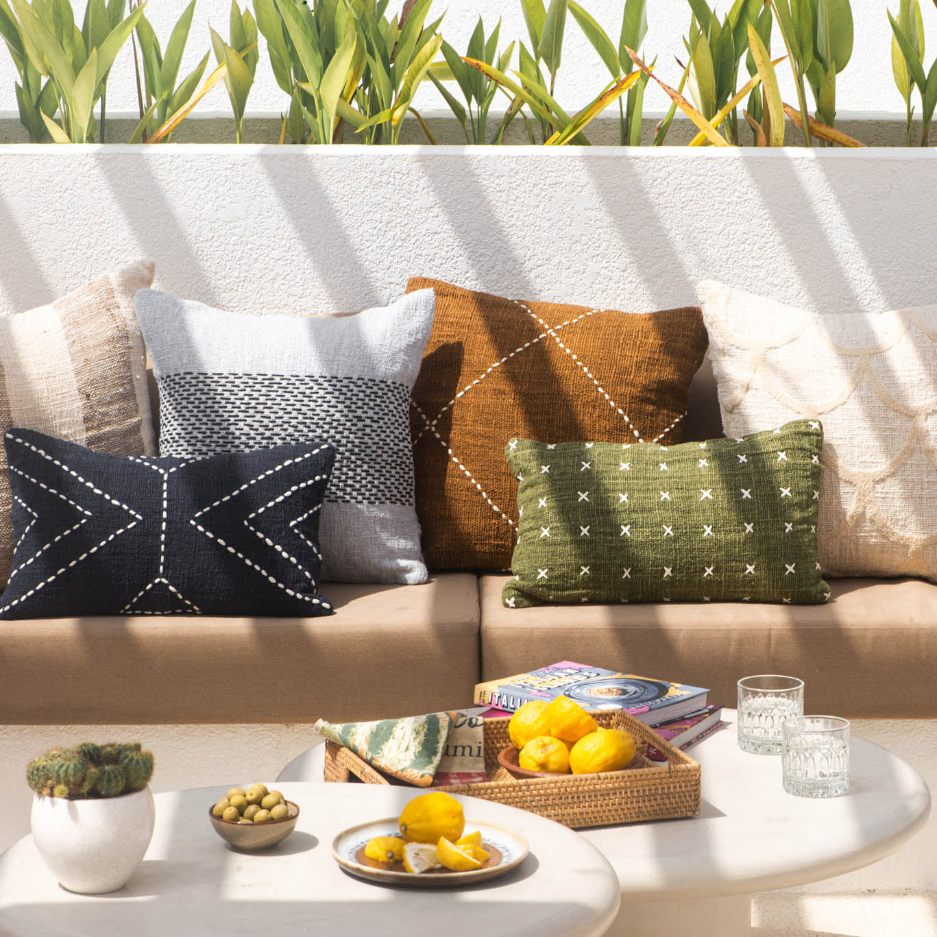 Outdoor seating area with a beige sectional sofa adorned with various throw pillows, a small table with fruit and glasses, and greenery in the background.