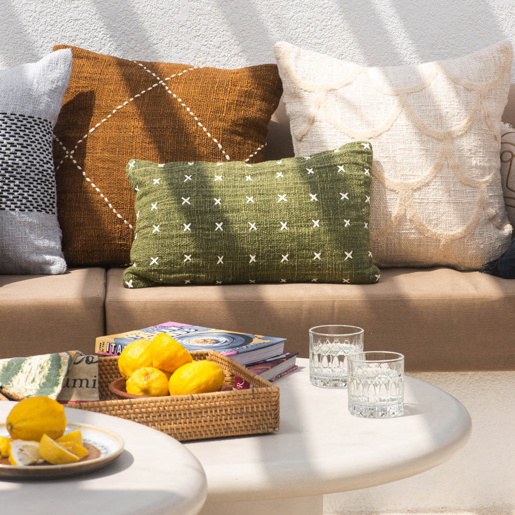 Outdoor setting with a beige sofa adorned with patterned cushions, a small table with lemons and glasses, and greenery in the background.